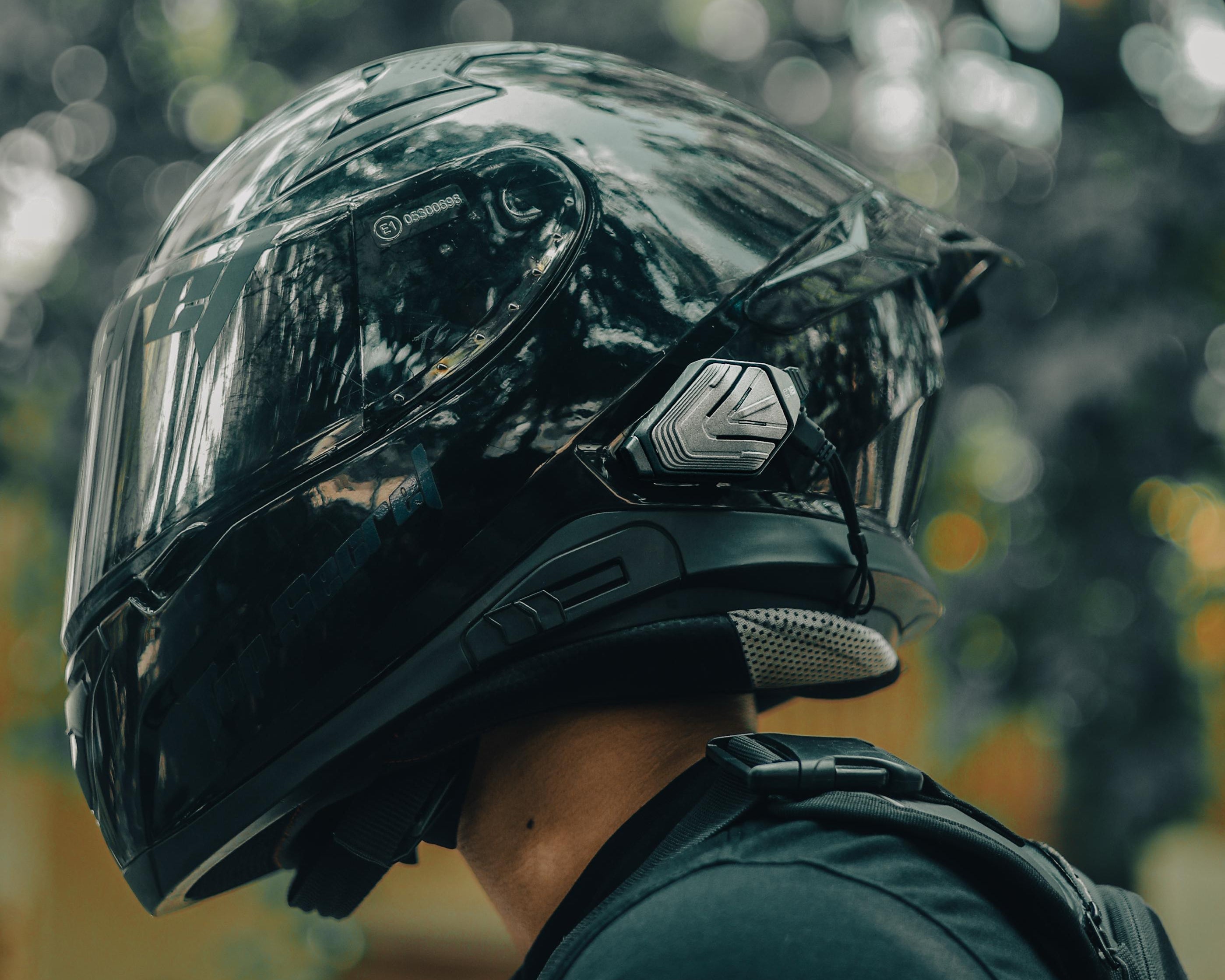 A close-up side profile of a person wearing a glossy black full-face motorcycle helmet with a clear visor and a dark shirt, photographed outdoors with a blurred natural background.