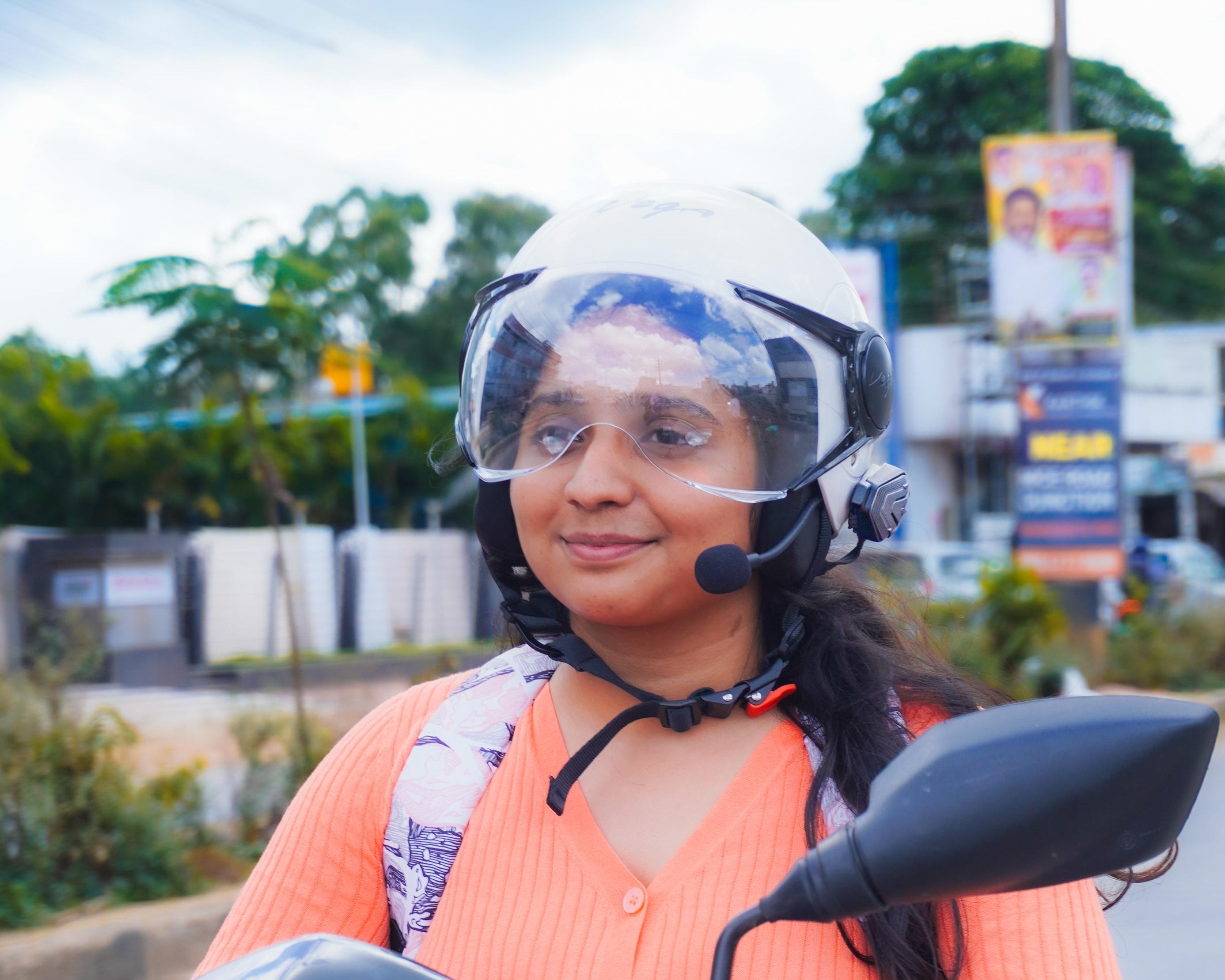 A woman wearing a white helmet and an orange top sits on a black scooter on a road with trees and buildings in the background.
